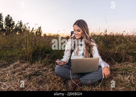 Young woman with a backpack on, using a laptop to charge his smartphone. Teleworking. Digital nomad Stock Photo