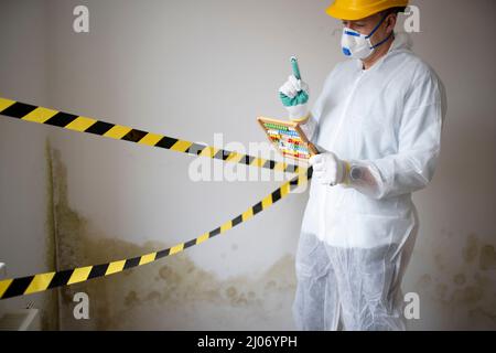 Man with white protective suit with abacus calculator, slide rule in hand and mouth nose mask in front of yellow black barrier tape in front of wall w Stock Photo