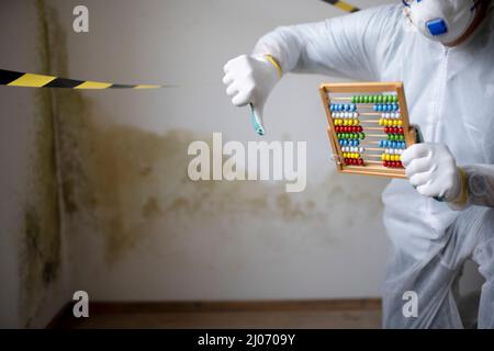 Man with white protective suit with abacus calculator, slide rule in hand and mouth nose mask in front of yellow black barrier tape in front of wall w Stock Photo