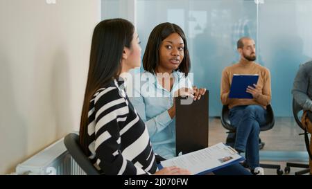 African american woman talking to asian candidate at job interview appointment. Female applicants waiting in queue and having conversation about hiring and recruitment, work application. Stock Photo