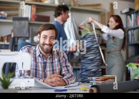 We can make anything. Portrait of a young fashion designer sitting in the workshop while his colleagues work in the background. Stock Photo