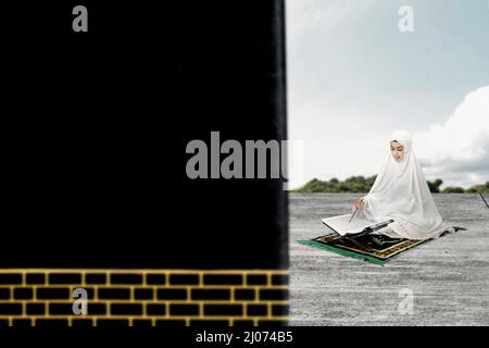 Asian Muslim woman in a veil sitting and reading the Quran in front of Kaaba with blue sky background Stock Photo