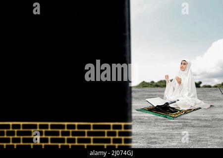 Asian Muslim woman in a veil sitting while raised hands and praying with prayer beads in front of Kaaba with blue sky background Stock Photo