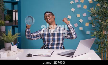 Positive adult holding clock and celebrating time passing, feeling happy about finished work before break. Young man looking at watch with hour and minutes, feeling excited about alarm. Stock Photo