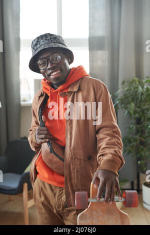 Vertical medium portrait of cheerful handsome cheerful African American man standing with skateboard in living room ready to go out smiling at camera Stock Photo