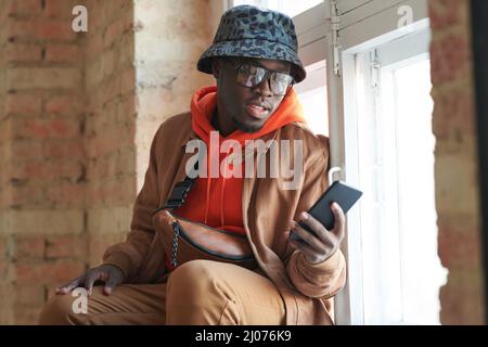 Horiozntal medium portrait of handsome young Black man wearing casual outfit and eyeglasses sitting on window sill having video call on smartphone Stock Photo