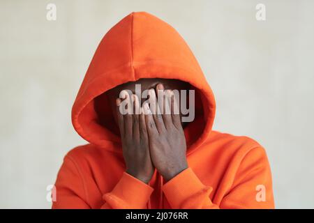 Close-up studio conceptual portrait shot of unrecognizable African American man wearing casual orange hoodie covering his face with hands Stock Photo