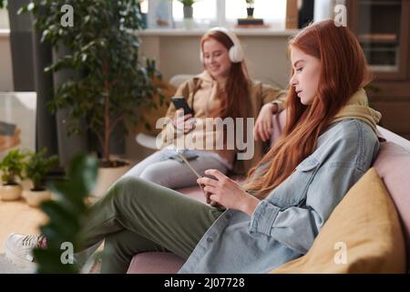 Side view portrait of young twin sisters wearing casual clothes spending time relaxing in living room at home surfing Interent and listening to music Stock Photo