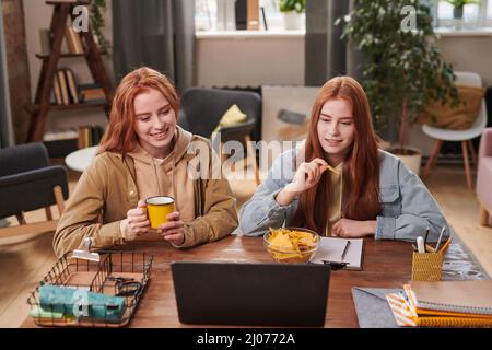 Horizontal high angle shot of modern young twin sisters spending time together at home watching series online on laptop and eating crisps Stock Photo