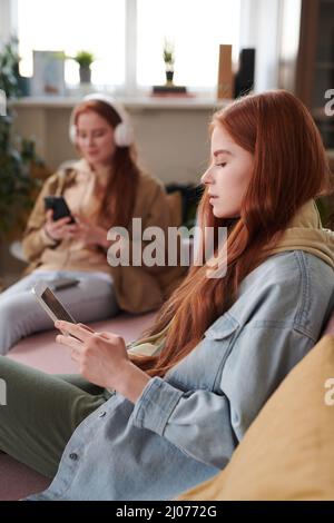 Vertical side view shot of modern twin sisters wearing casual clothes spending time relaxing in living room at home using gadgets Stock Photo
