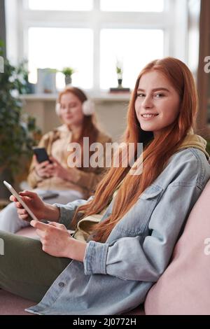 Caucasian twin sisters wearing casual outfits spending spare time relaxing in living room at home using gadgets, one of them looking at camera Stock Photo