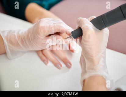 Manicurist using cuticle remover device while doing hardware manicure in beauty salon. Close up of female manicure master in sterile gloves removing cuticle on woman hand with electric nail drill. Stock Photo