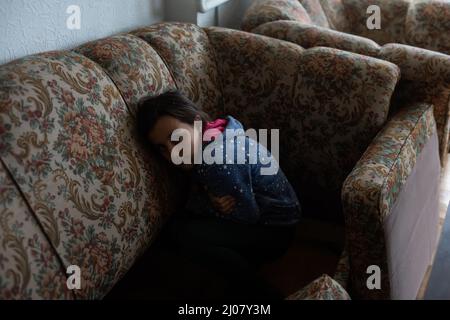 little Ukrainian girl is hiding in the sofa, looks confused Stock Photo