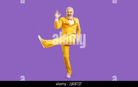 Happy funny senior man in yellow party suit dancing isolated on purple background Stock Photo