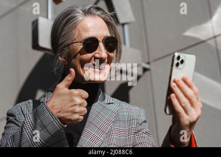 Grey white senior woman in sunglasses gesturing while using cellphone outdoors Stock Photo