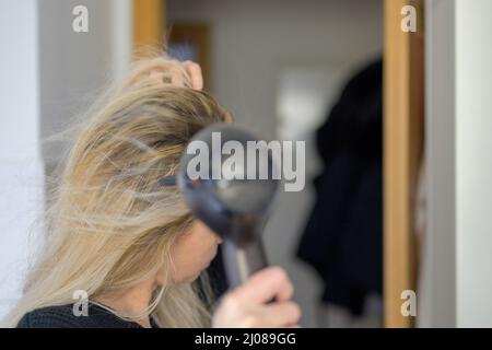 Woman drying her long blond hair with a hairdryer after washing it in a closeup side view Stock Photo