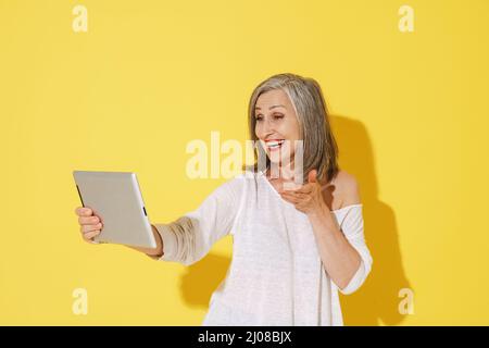 White senior woman gesturing while using tablet computer isolated over yellow wall Stock Photo
