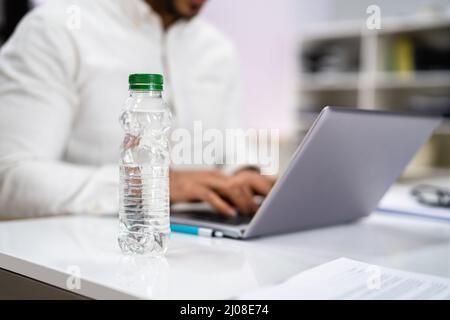 Water Bottle On Desk And Man In Foreground Using Computer Stock Photo