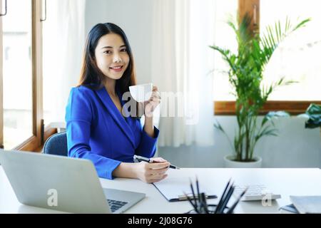 Manager, entrepreneur, female employee sitting and drinking coffee expressing a smiling face while taking a break from hard work. Stock Photo