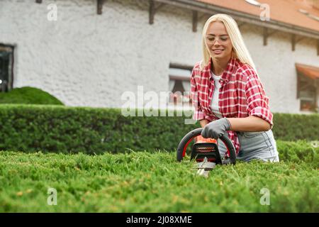 Positive young woman with blond hair wearing safety glasses and gloves using electric trimmer for taking care of bushes outdoors. Seasonal work at garden. Stock Photo