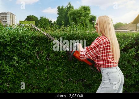 Side view of attractive caucasian woman in casual wear pruning hedge with modern gardening tool. Pleasant female with blond hair enjoying working process at garden.  Stock Photo