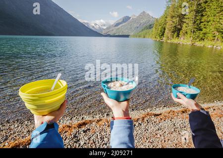 Friends on a hike eat porridge for breakfast in tourist plates and bowls. Against the background of an idyllic landscape of a mountain lake. Food and Stock Photo