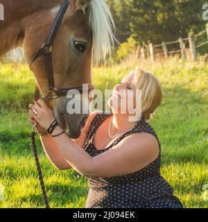 Blonde pretty young woman with Haflinger horse. The woman sits in front of the horse and smiles happily. Outside in the pasture with backlight in summ Stock Photo