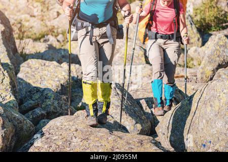 Two hikers wearing waterproof gaiters over trekking boots walking with hiking sticks among boulders in nature park. Clothing and equipment for backpac Stock Photo