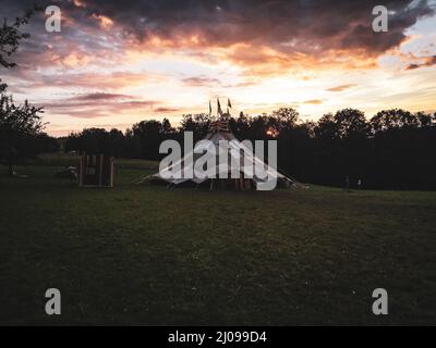Beautiful view of a cloth tent on a green grass and silhouetted trees against a sunset orange sky Stock Photo