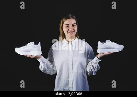 A fair-haired woman in a white shirt on a black background holds white sneakers made of genuine leather in her hands Stock Photo