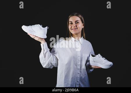 A fair-haired woman in a white shirt on a black background holds white sneakers made of genuine leather in her hands Stock Photo