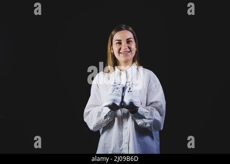 A fair-haired woman in a white shirt on a black background holds white sneakers made of genuine leather in her hands Stock Photo