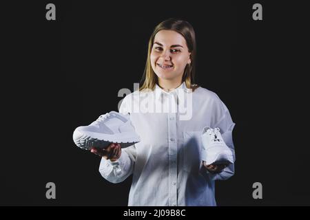 A fair-haired woman in a white shirt on a black background holds white sneakers made of genuine leather in her hands Stock Photo