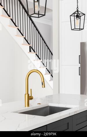 A kitchen sink detail shot with a gold faucet, grey island, white marble countertop, and lights hanging above the island. Stock Photo