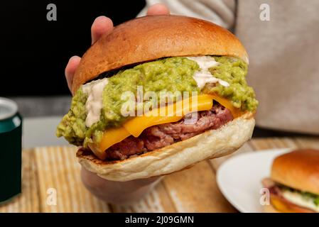 A delicious double cheddar beef patty with lots of guacamole and mayonnaise on top of the meat on a lightly toasted bun held in a person's hand on a w Stock Photo