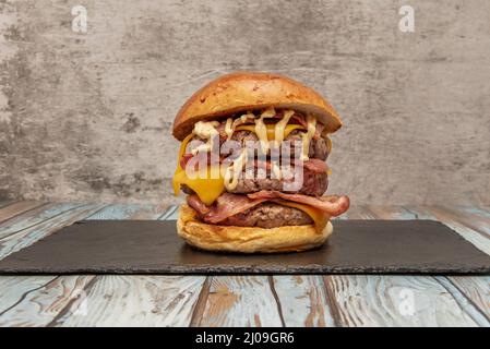 a beastly three meat burger with lots of fried bacon, cheddar cheese and sauce to give you a heart attack and a good bite on a black slate board and b Stock Photo