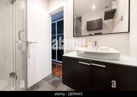 Bathroom with glass-enclosed shower stall, angular white porcelain sink on glass top, and dark wood wall unit with black-framed mirror Stock Photo