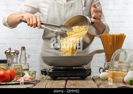 Chef in white uniform pours boiled spaghetti into frying pan. Backstage of cooking pasta alla carbonara. Traditional italian dish on white background. Stock Photo