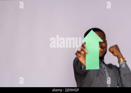 African man holding a green arrow pointing upwards and covering his face on a gray background Stock Photo