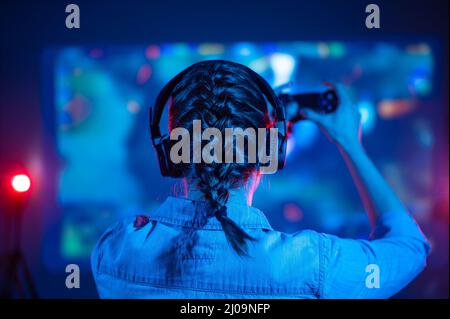 View from back of young emotional gamer woman with pigtail playing video game at home in front of big screen with headphone and joystick. Colorful neo Stock Photo