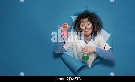 Foolish crazy chemist doing silly grimace and holding beakers after insane chemical explosion. Mad goofy chemist having messy hair and funny amusing facial expressions in studio. Stock Photo