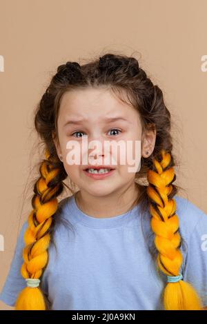 Upset little girl with open mouth and tears in eyes crying having kanekalon braids of yellow colour on head wearing light blue t-shirt on beige Stock Photo