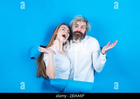 Surprised couple of bearded man and happy woman making hole in paper looking through hole. Advertising board, blue background. Couple through hole in Stock Photo