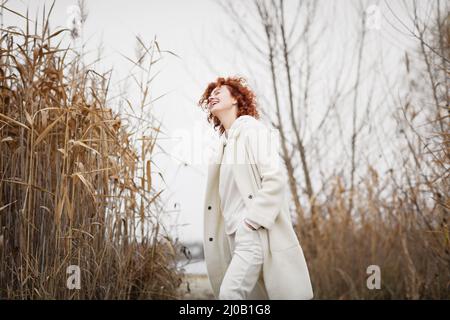 Beautiful young woman in trendy white clothes poses in motion and laughing against the backdrop of an autumn landscape with dry reeds Stock Photo