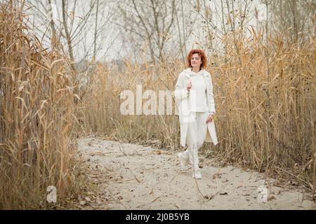 Pretty young woman in trendy white clothes poses in motion against the backdrop of an autumn landscape with dry reeds Stock Photo