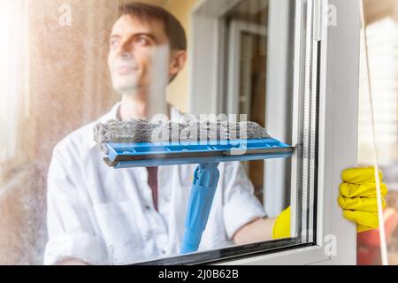 Cleaning concept. Smiling young man washing window in yellow gloves using squeegee or rag, close up. Spray detergent. Cleaning dirty windows, home hyg Stock Photo