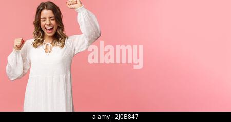 Portrait of beautiful tender young blond girl in white dress, raising hands up in hooray, yes or victory, shouting relieved, scream from happiness Stock Photo