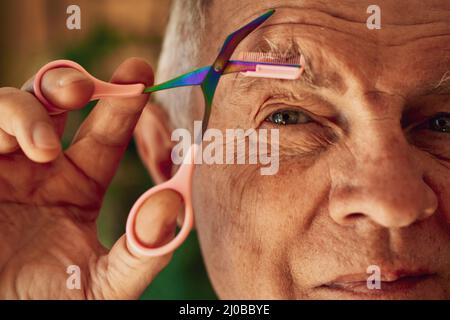 Eyebrow trimmer. Adult mature man trims his eyebrow hair with scissors with a comb. Facial hair removal Stock Photo