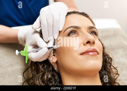 Crop beautician in gloves doing filler injection in cheek zone to charming female client during rejuvenation procedure in cosmetology clinic Stock Photo