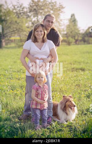 Pregnant Woman and Her Husband with her daughter holding her hands in a heart shape on her baby Stock Photo